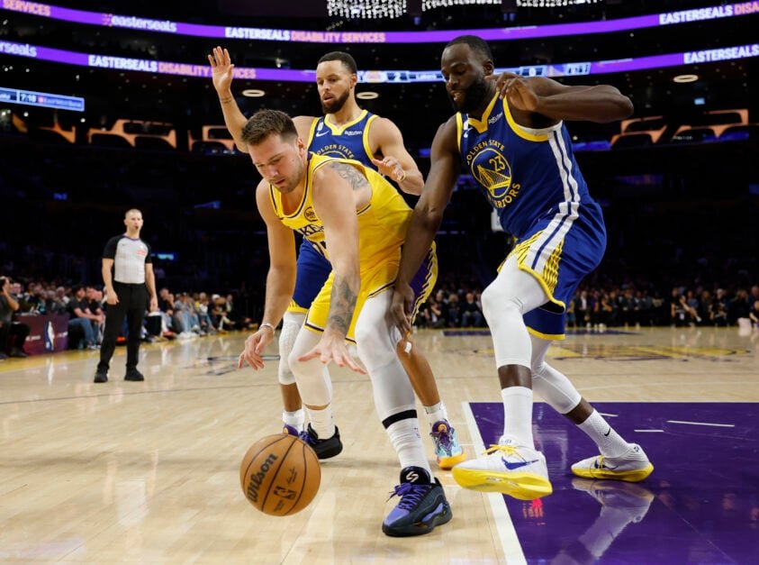 Luka Doncic #77 of the Los Angeles Lakers reaches for the ball while being protected by Draymond Green #23 and Stephen Curry #30 of the Golden State Warriors